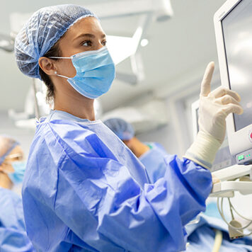 Portrait of a young female doctor in scrubs and a protective face mask preparing an anesthesia machine before an operation.